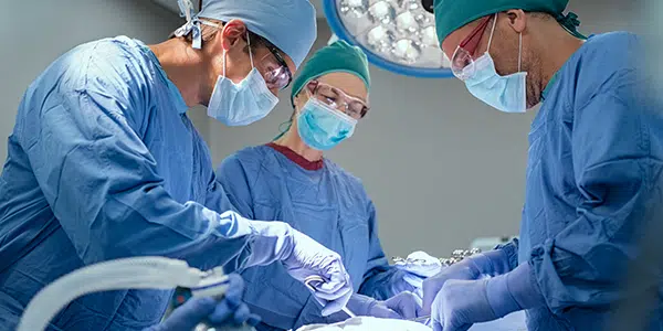 Três profissionais de saúde em sala de cirurgia, operando paciente, seguindo as normas de metas internacionais de segurança do paciente.