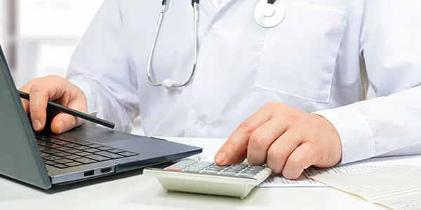 Médico digitando em uma calculadora e fazendo anotações no computador